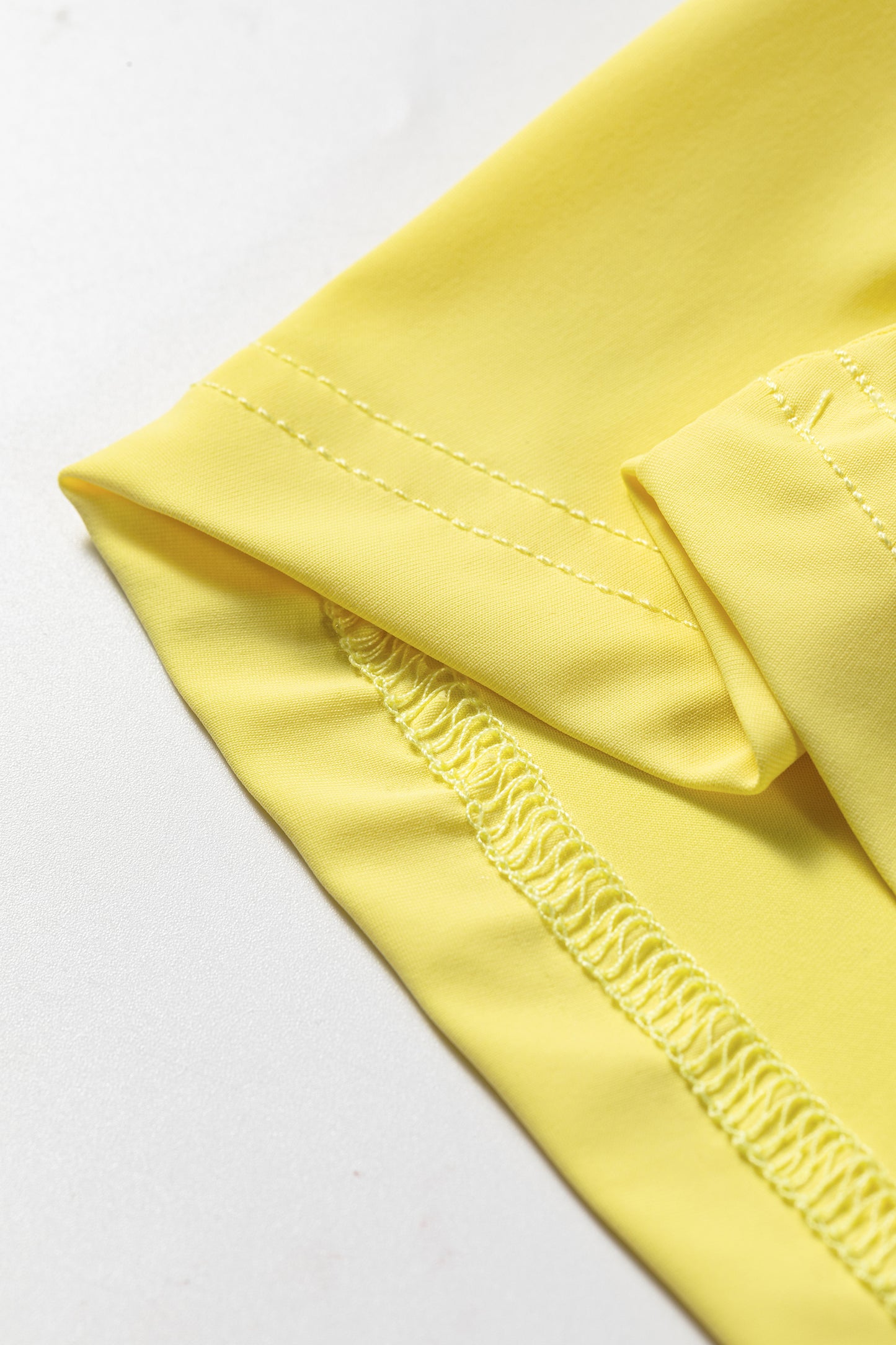 A close-up of a light yellow fabric garment, showing the texture and double-stitched hem detail on a white background.