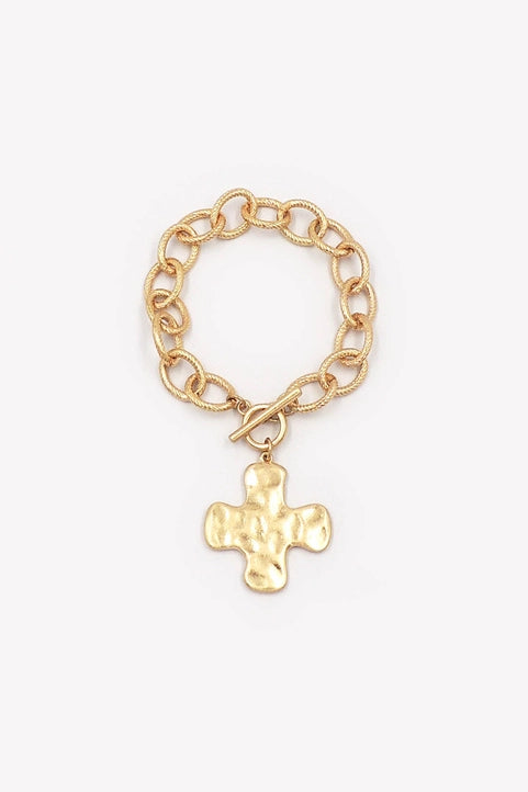 Worn gold link toggle bracelet by What's Hot featuring a hammered cross charm, showcasing layered jewelry style.