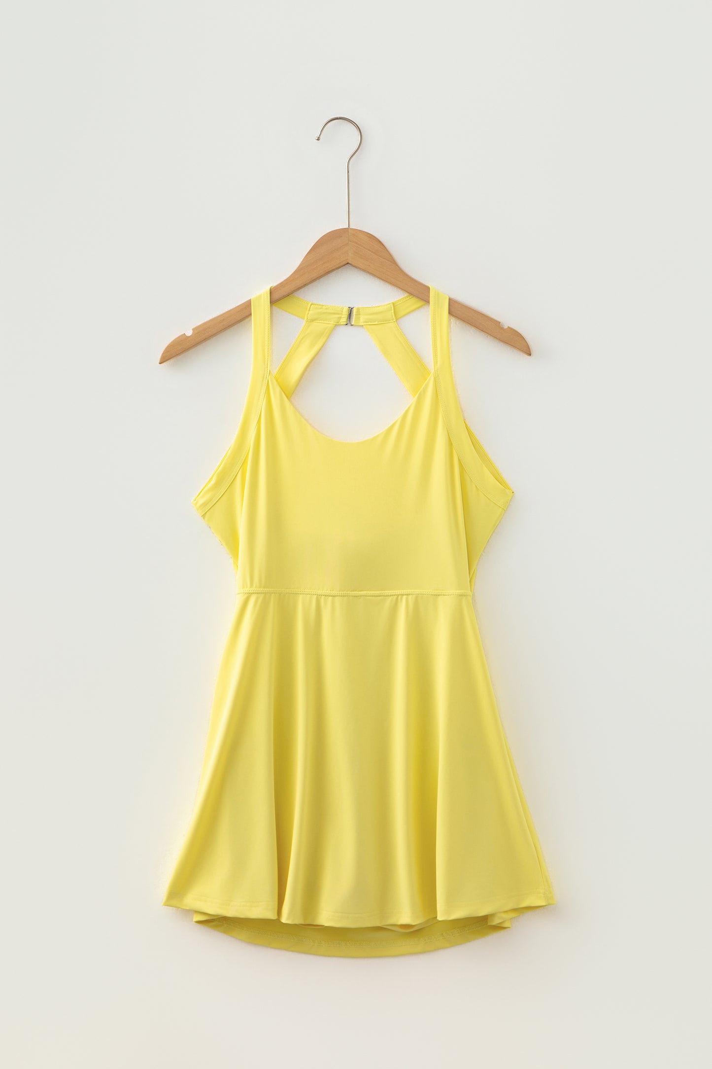 A bright yellow sleeveless active dress with a crisscross cutout back, hanging on a wooden hanger against a plain off-white wall.