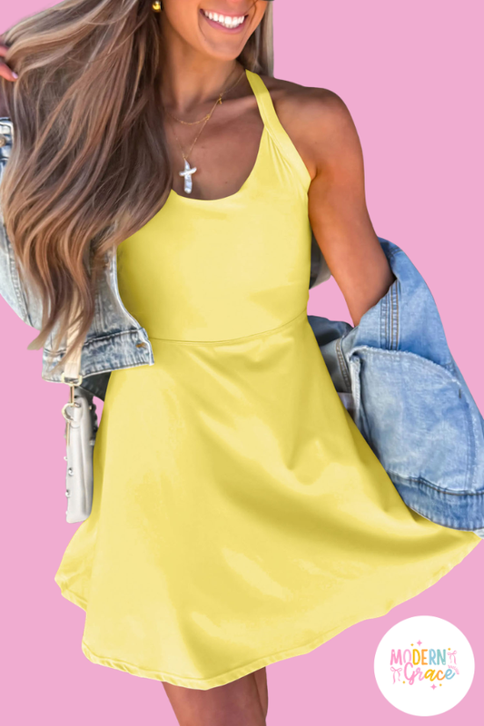 A woman wearing a bright yellow sleeveless fit-and-flare mini active dress, styled with a light blue denim jacket draped over her shoulders, set against a solid pink background.