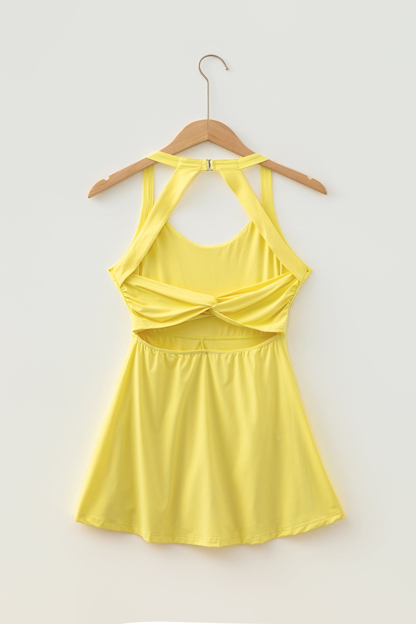 A bright yellow halter-neck active dress with a twisted bust detail and a flared skirt, hanging on a wooden hanger against a plain light gray background.